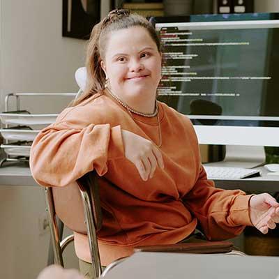 A person sits in an office chair wearing a loose orange sweatshirt, with a computer monitor displaying lines of code in the background.