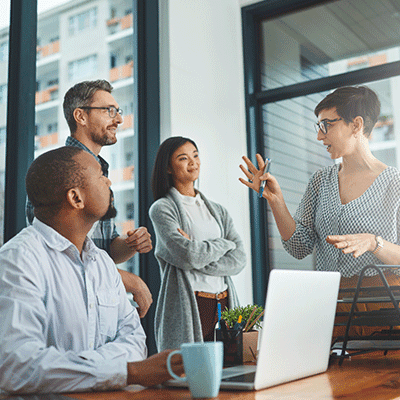 Four diverse professionals collaborating in a modern office meeting, with a woman gesturing while presenting ideas to colleagues.