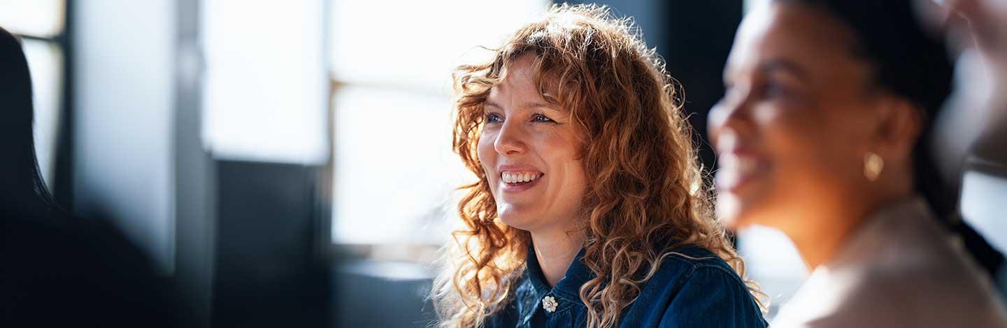 Woman with curly brown hair smiling in an office setting with a colleague beside her in a bright, professional environment.