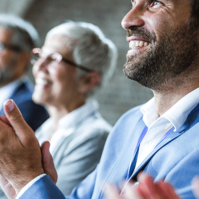 Smiling bearded man in blue shirt clapping with blurred diverse colleagues in background during business meeting