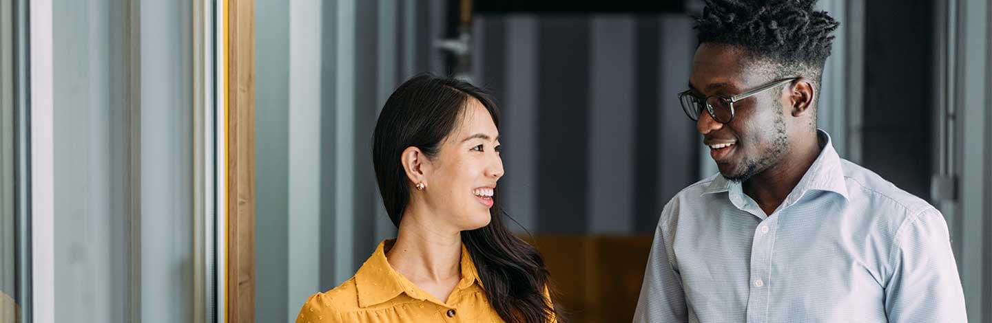 Two people walk together in a modern office hallway, engaged in conversation. One holds a tablet while gesturing with their other hand, and the other listens attentively with hands loosely clasped.