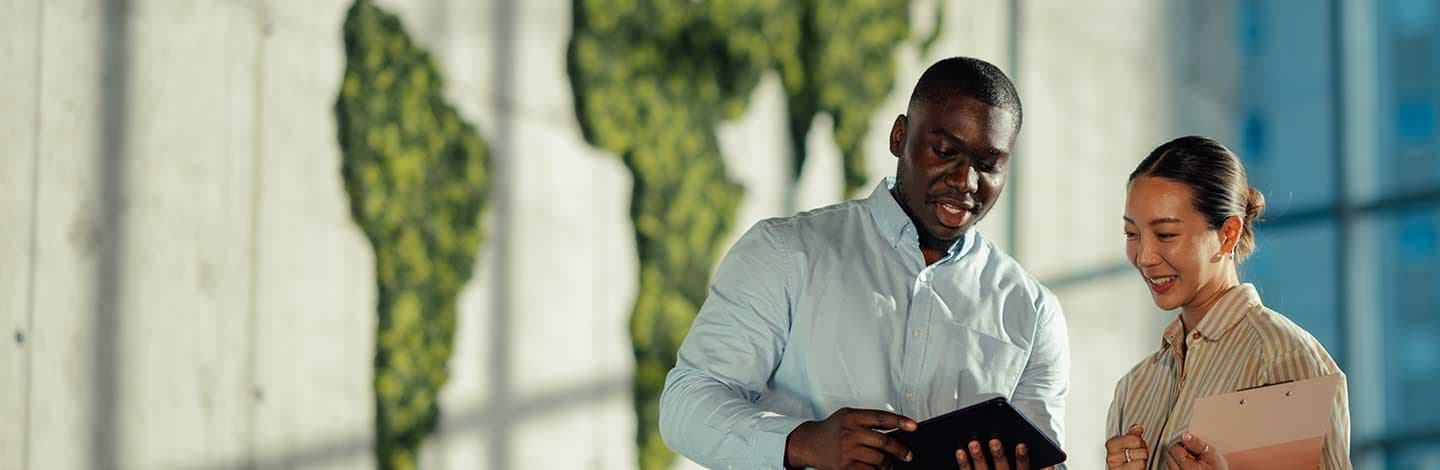 Two colleagues, standing in a modern office setting with a green foliage world map on the wall behind them. The man holds a tablet and gestures while speaking, and the woman smiles, holding a pink folder.