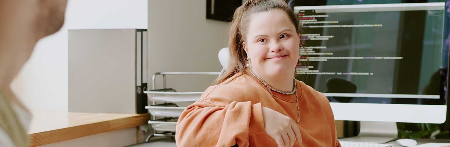 A person sits in an office chair wearing a loose orange sweatshirt, with a computer monitor displaying lines of code in the background.