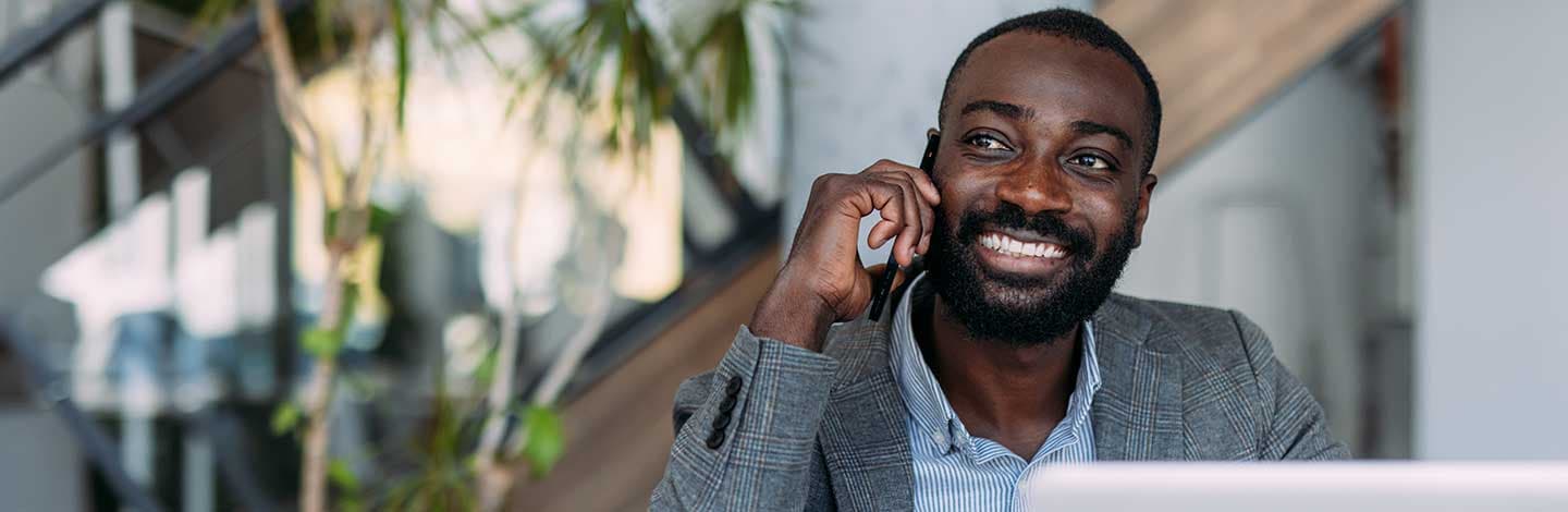 A person sits at a desk in a modern office, speaking on a mobile phone while working on a laptop. A staircase and indoor plants are visible in the background.