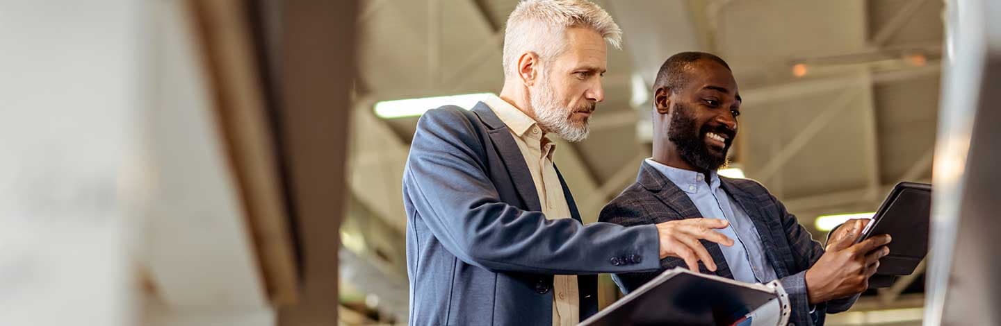 Two businessmen in suits collaborating while looking at a tablet computer in a modern office setting.