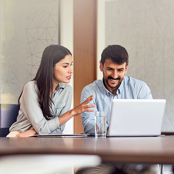 Two business professionals collaborating at a laptop in a modern office, woman gesturing while man smiles at screen.